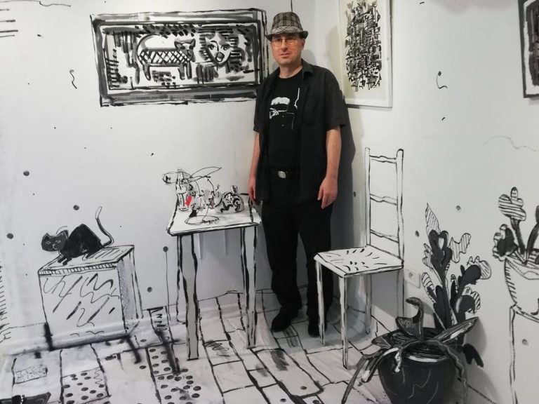 Homme en noir se tenant dans une pièce peinte en monochrome avec des objets esquissés.